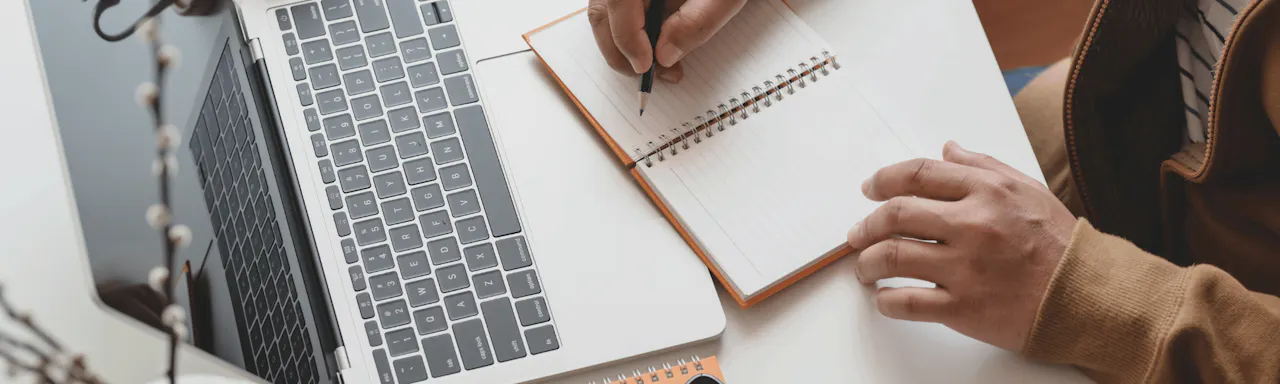 Person Writing on Notebook next to Laptop